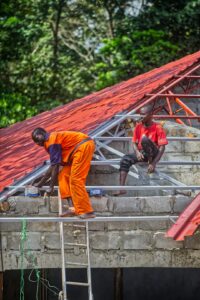 Workers inspect a protected residential roof beside ladders, covered materials, gutters, and renovation tools.